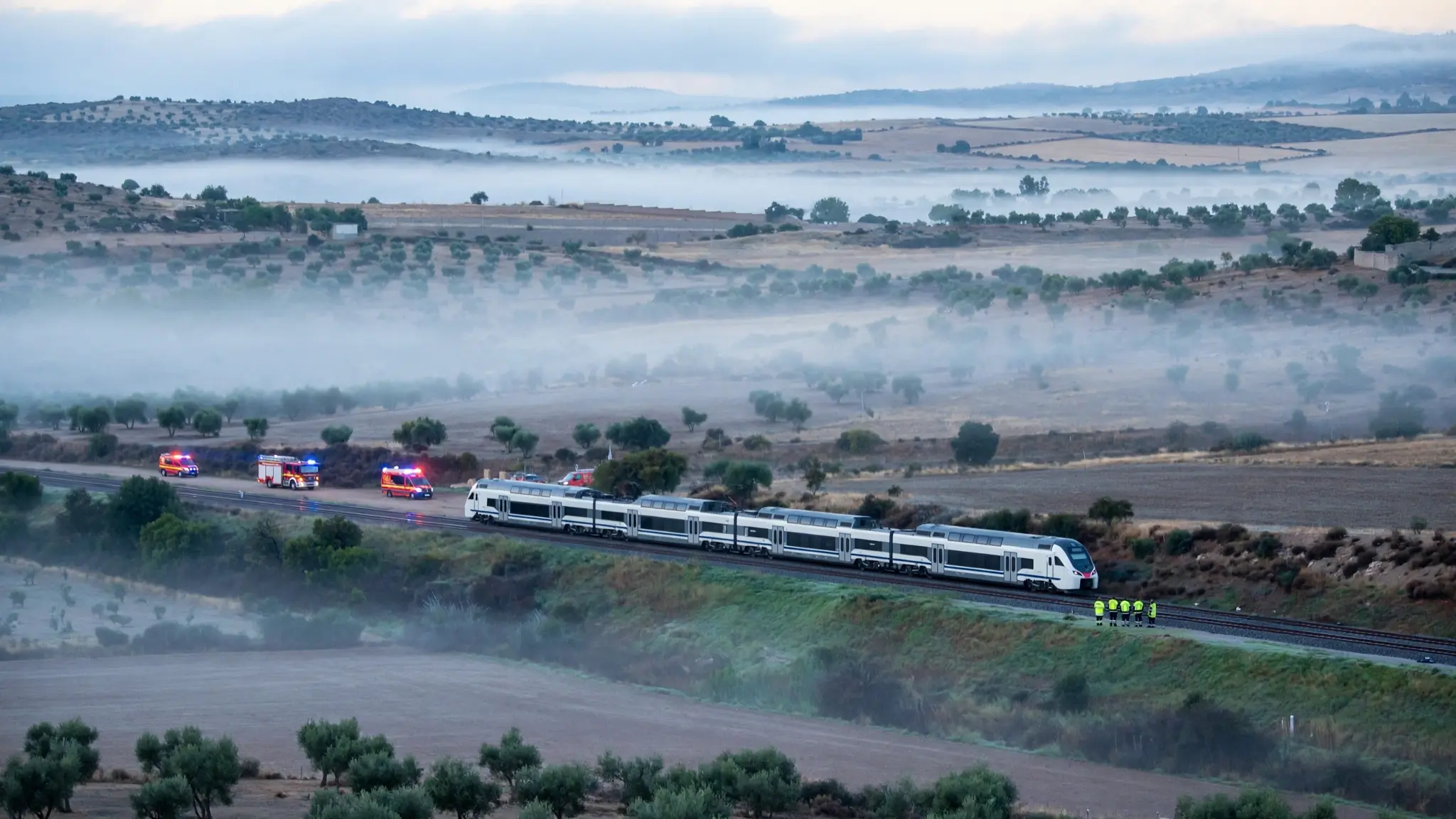 Un tren de pasajeros a lo lejos en el campo de Toledo al amanecer, con vehículos de emergencia y personal con chalecos reflectantes a distancia, sin personas identificables