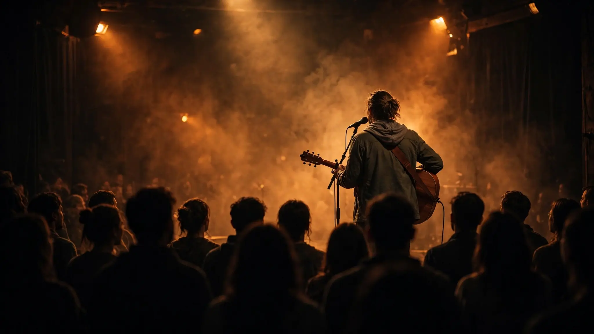 A singer-songwriter seen from behind at a microphone on a dimly lit stage, holding an acoustic guitar in warm amber light